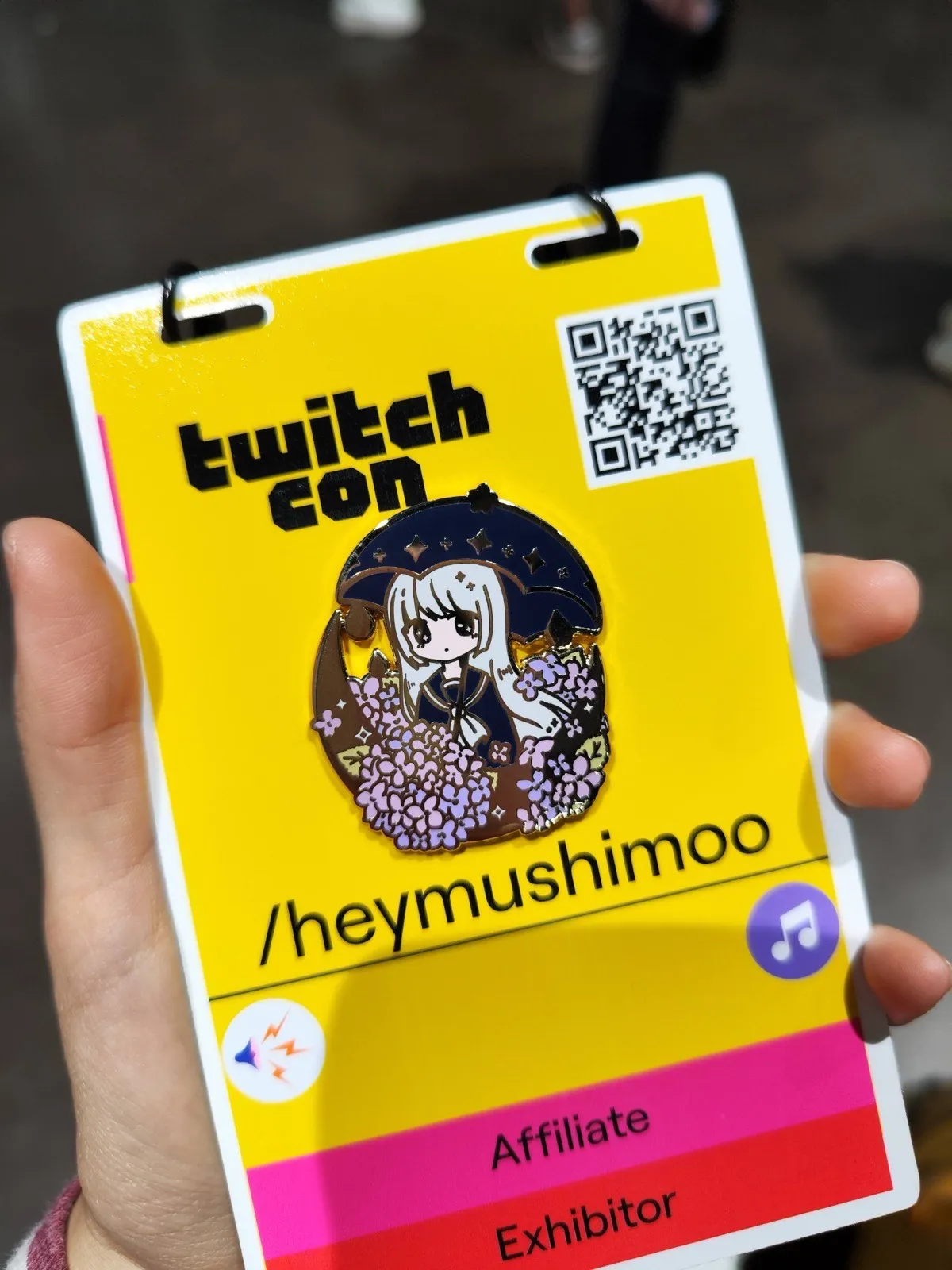 A hand holding a yellow TwitchCon exhibitor badge reading slash heymushimoo, pinned with an enamel pin of a white-haired character under an umbrella among purple hydrangeas