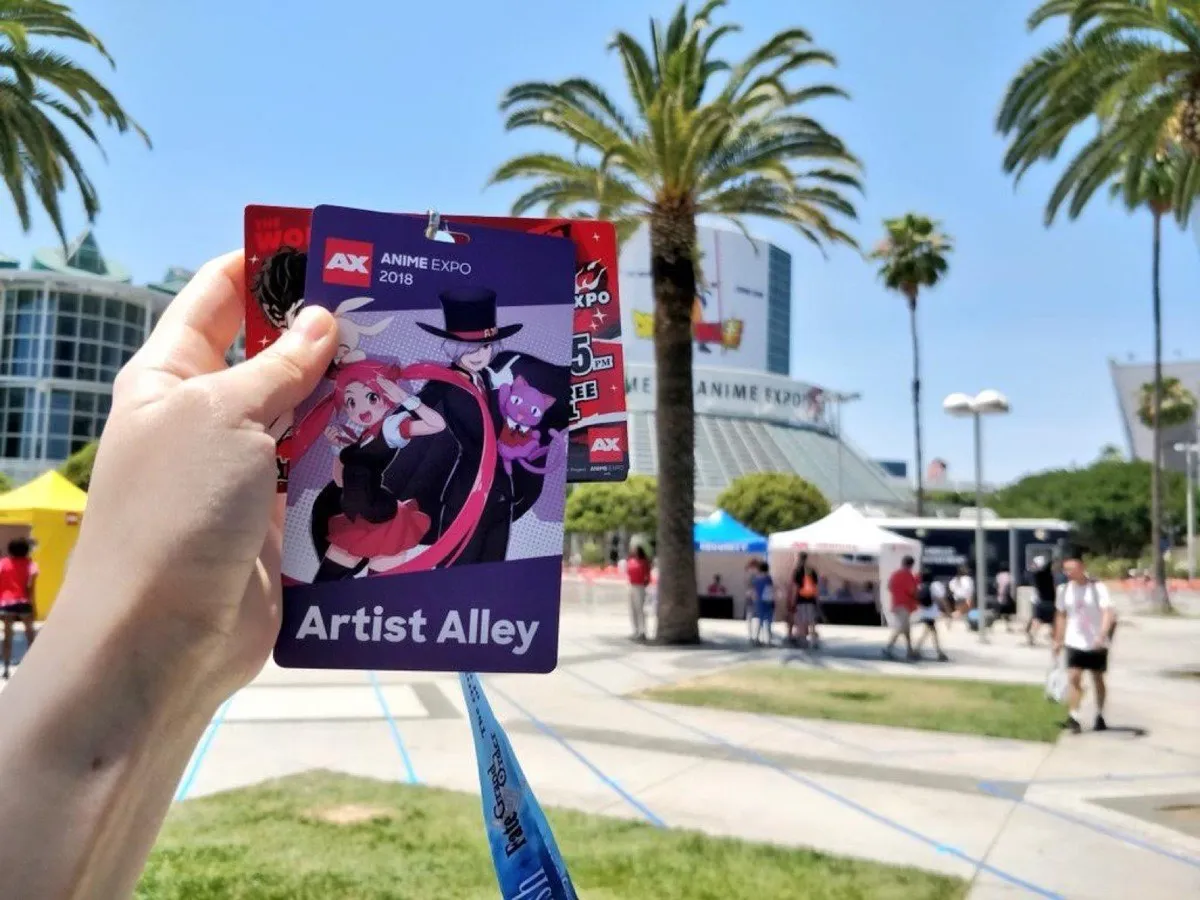 A hand holding an Anime Expo 2018 Artist Alley badge in front of the Los Angeles Convention Center, palm trees and an Anime Expo banner behind it