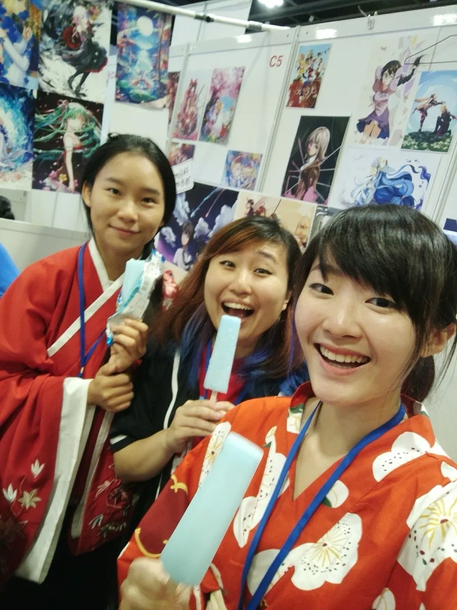 Selfie of Xinni and two friends in yukata at artist alley booth C5, holding pale blue ice lollies, a wall of anime prints behind them