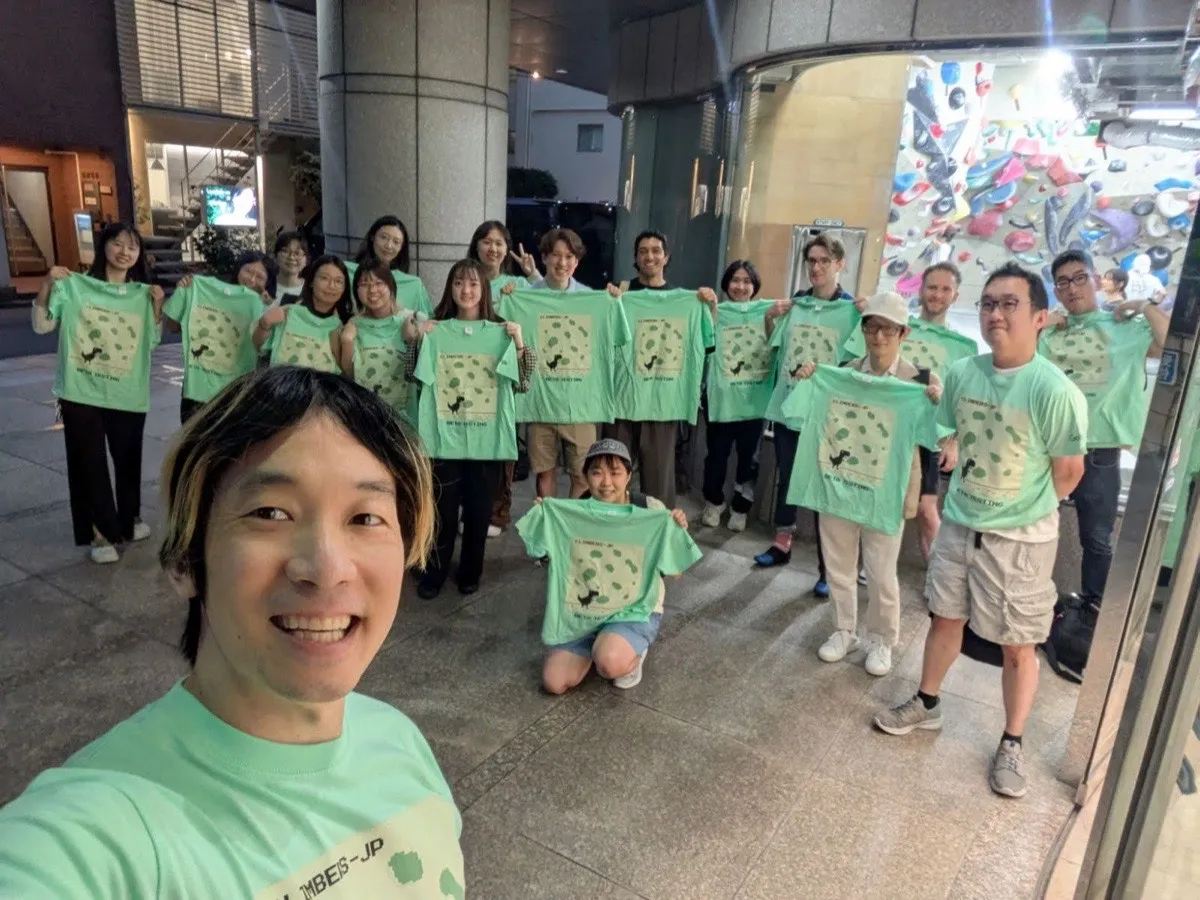 About eighteen people outside a bouldering gym at dusk, each holding up a mint-green tee printed with a small dark animal at the foot of a wall of green climbing holds and the words CLIMBERS-JP
