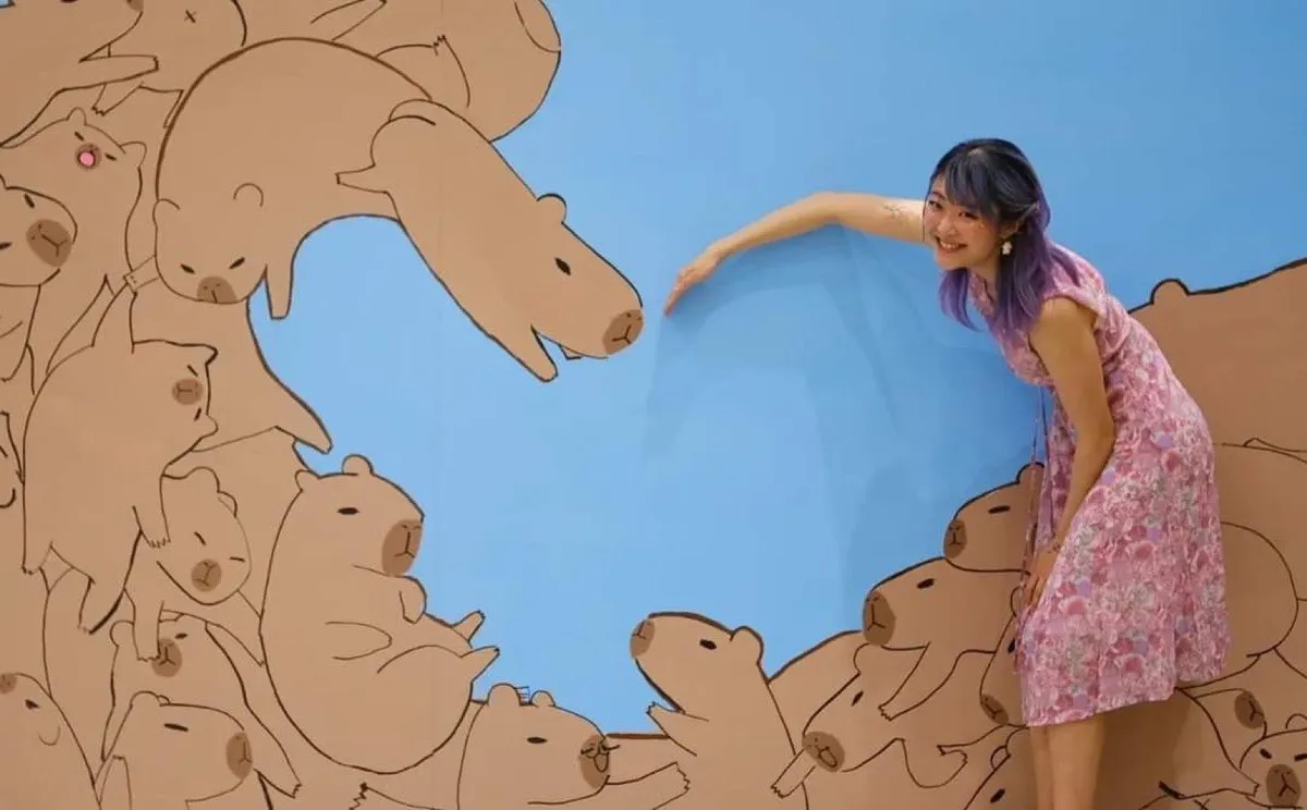Xinni leaning in from the right of a wall covered in line-drawn capybaras piled around a heart-shaped gap of blue, touching the nose of the largest one