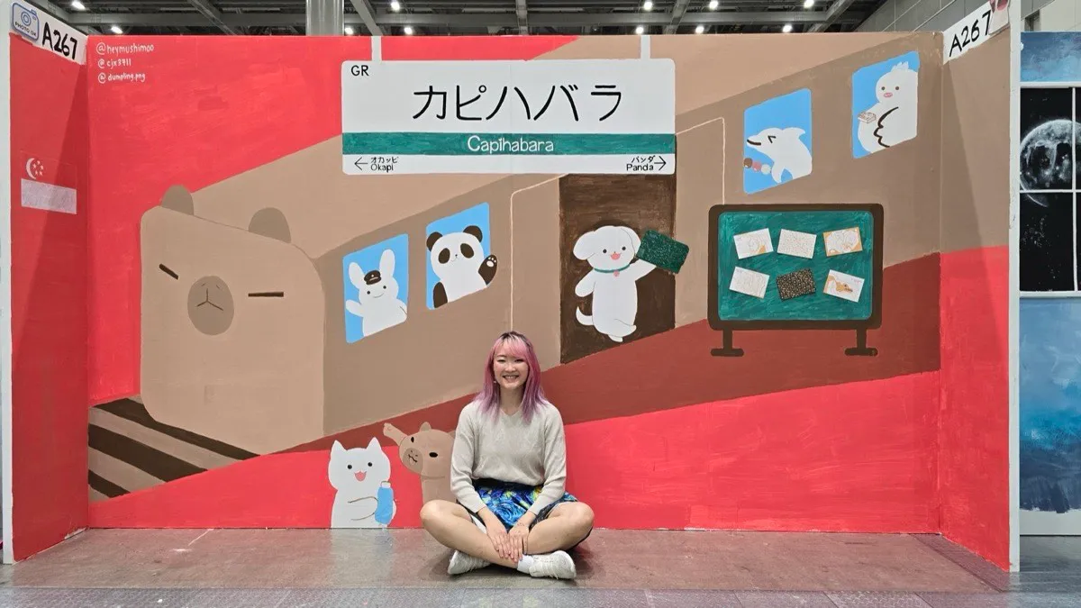 Xinni sitting cross-legged in front of a hand-painted booth mural of a train station named Capihabara, with a capybara-fronted train, a rabbit in a station-master cap, a panda and a white dog holding a sign