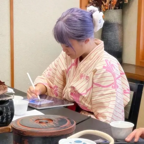 Xinni in a cream kimono with purple hair drawing on an iPad with an Apple Pencil at a low lacquer table