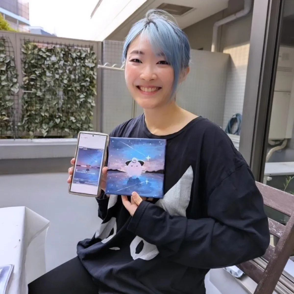 Xinni holding up a small square canvas she painted of a round pink character standing in shallow water under a starry sky, next to her phone showing the digital original