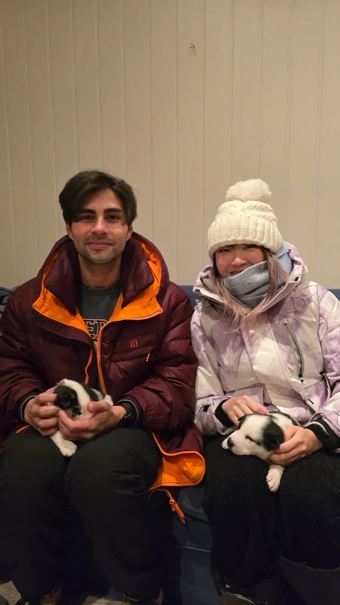 Xinni in a bobble hat and ski jacket sitting beside a friend in a puffer jacket, each holding a small black-and-white puppy