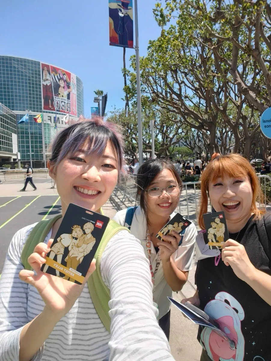 Selfie of three of us outside the Los Angeles Convention Center holding up Anime Expo 2022 Artist Alley badges, a One Piece Film Red billboard on the building behind