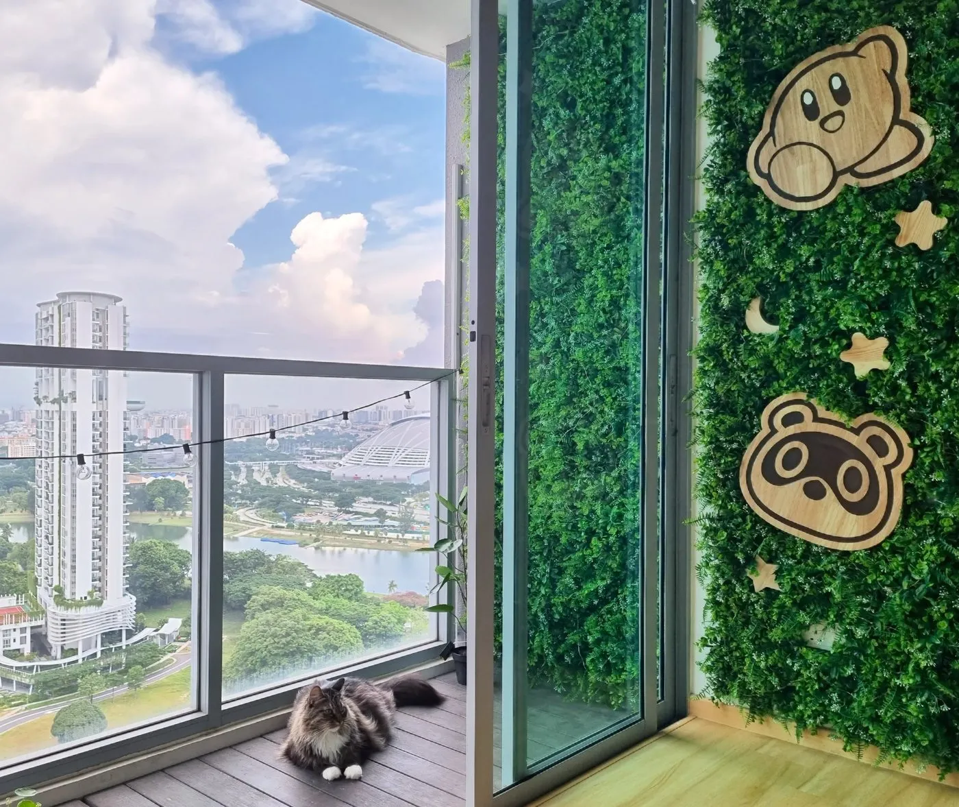 A high-rise balcony with a green wall bearing wooden Kirby and Animal Crossing cut-outs, festoon lights, a long-haired cat resting on the deck, and a Singapore park-and-city view beyond the glass