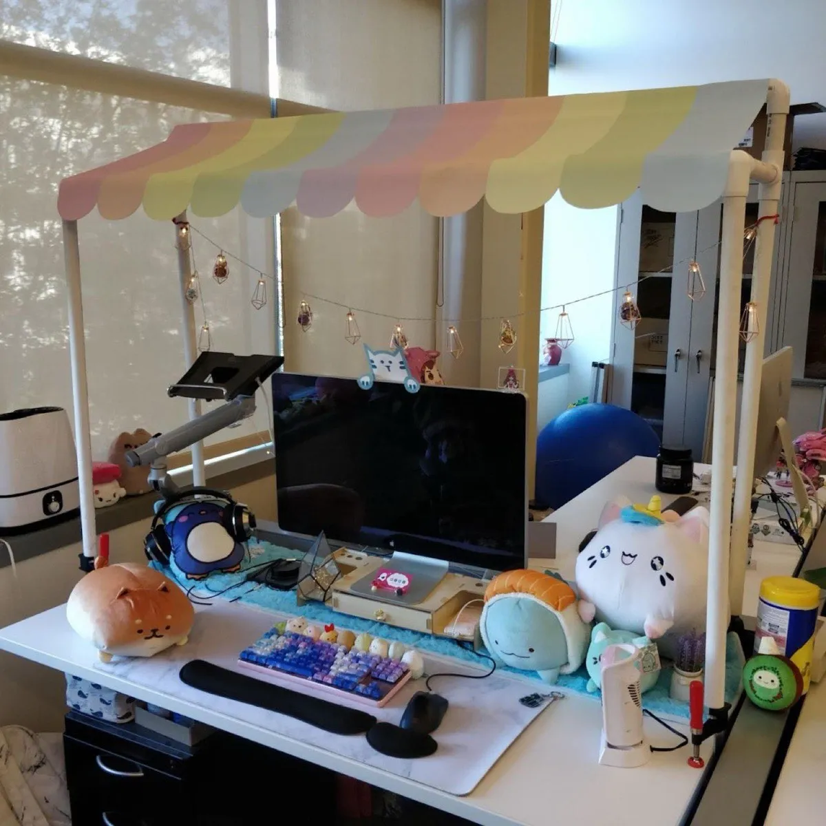 An office desk built out as a market stall: a pastel scalloped awning on a pipe frame, fairy lights, a monitor, a pastel mechanical keyboard with novelty keycaps, and plush toys crowded around it