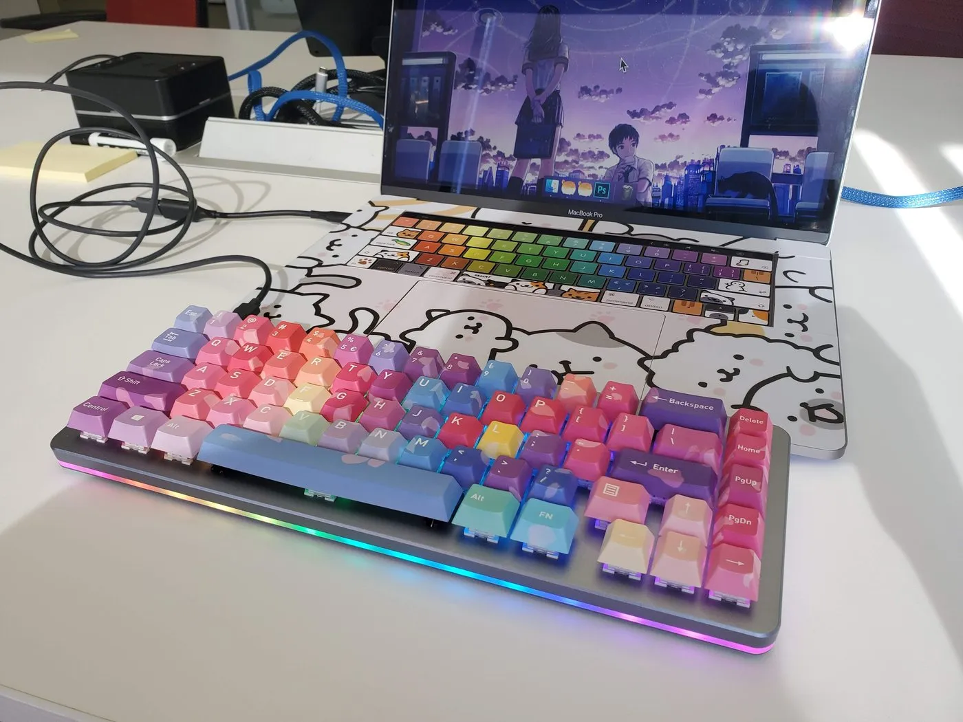 A custom mechanical keyboard with rainbow gradient keycaps and RGB underglow, in front of a MacBook with a Shinkai wallpaper and a cat-illustrated keyboard cover