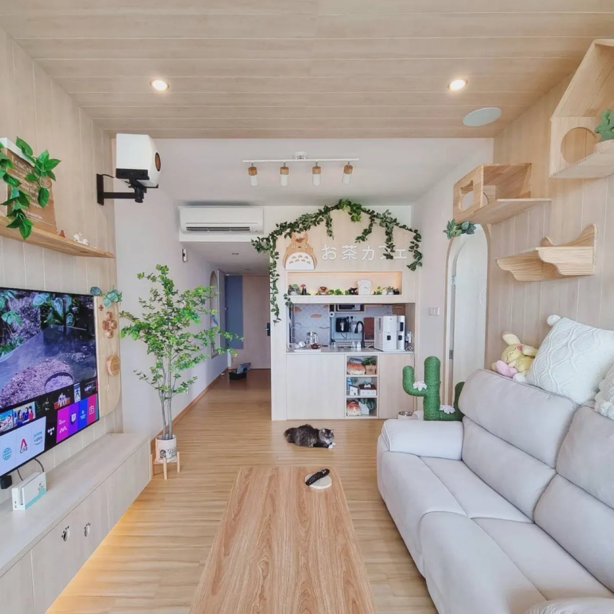 A pale wood-panelled living room: a wall-mounted TV, a tea nook under a Totoro cut-out and a sign reading お茶カフェ, cactus-shaped cat scratching posts, wall-mounted cat shelves, plush toys on a cream sofa, and a long-haired grey cat lying on the floor