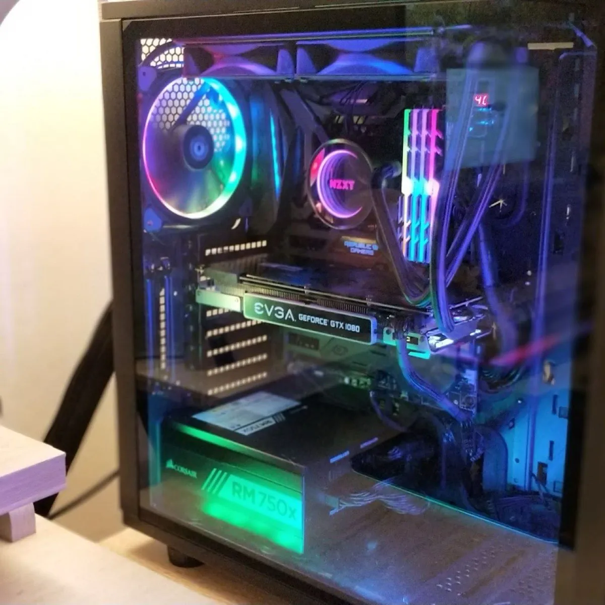 A glass-sided PC build lit in RGB on a desk: a large ring-lit intake fan, an NZXT pump, lit RAM, an EVGA GeForce GTX 1080 and a Corsair RM750x power supply