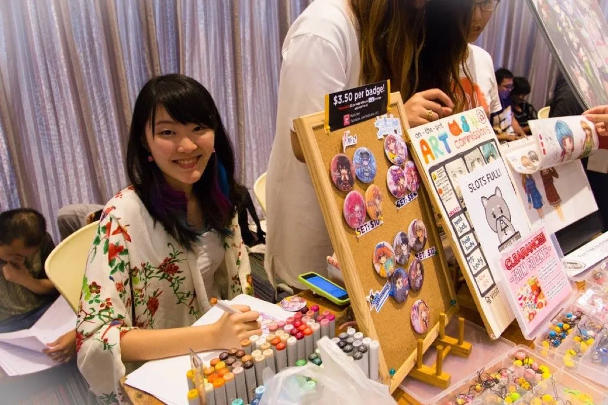 Xinni behind the mushimoo booth at an artist alley, badges racked up beside her