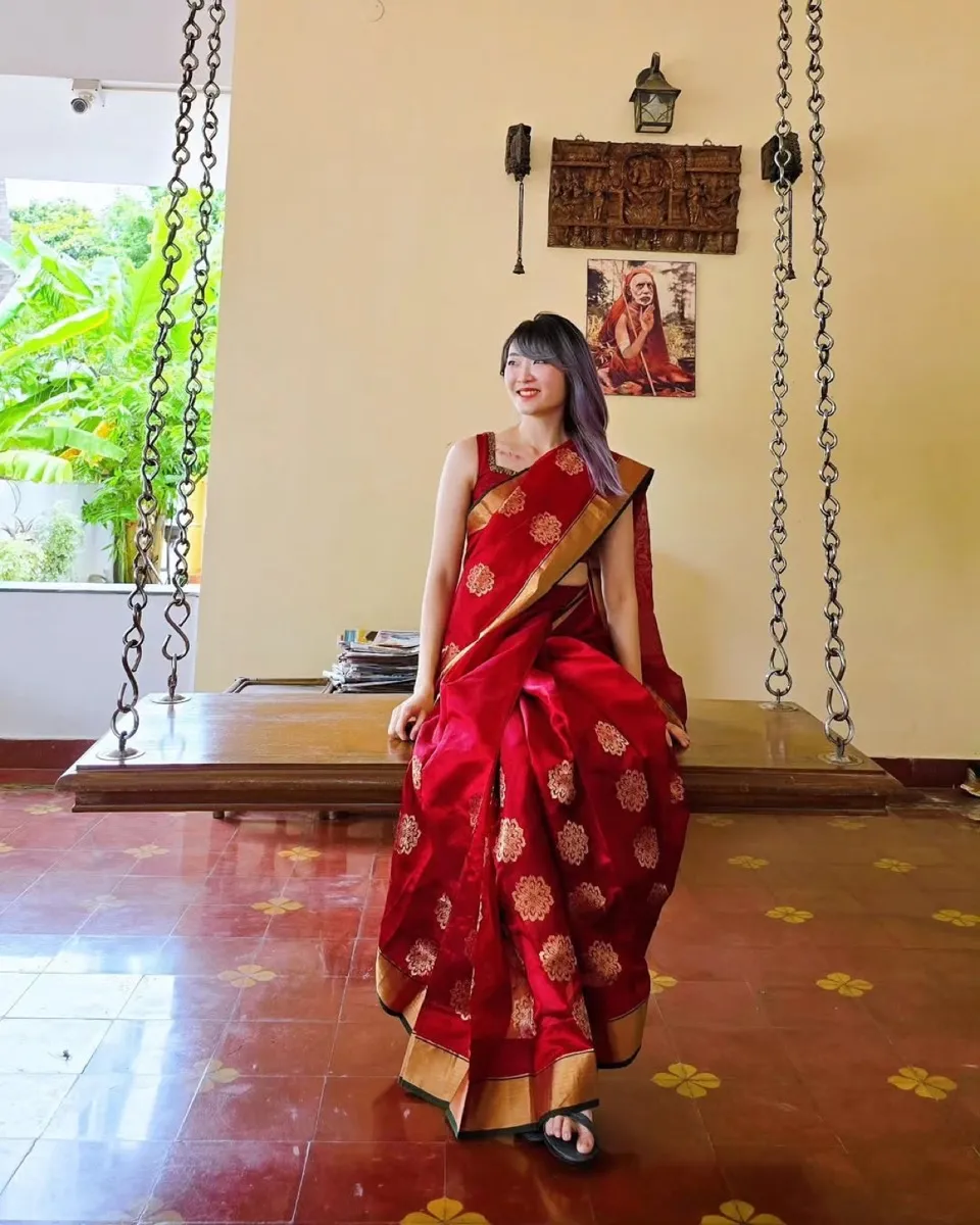 Xinni in a red and gold silk saree, leaning against a wooden swing seat in a tiled room with greenery through the window behind her