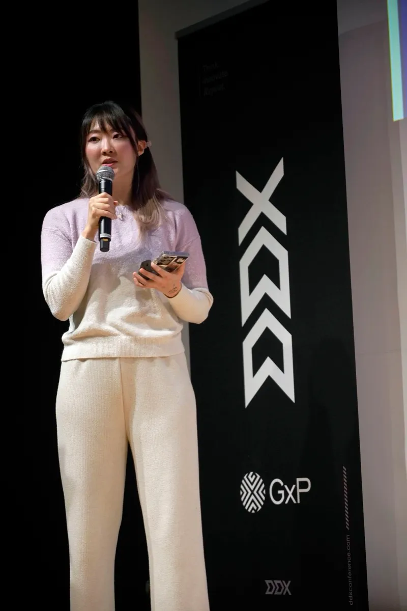 Xinni on stage holding a microphone and a phone, speaking in front of a tall black DDX conference banner