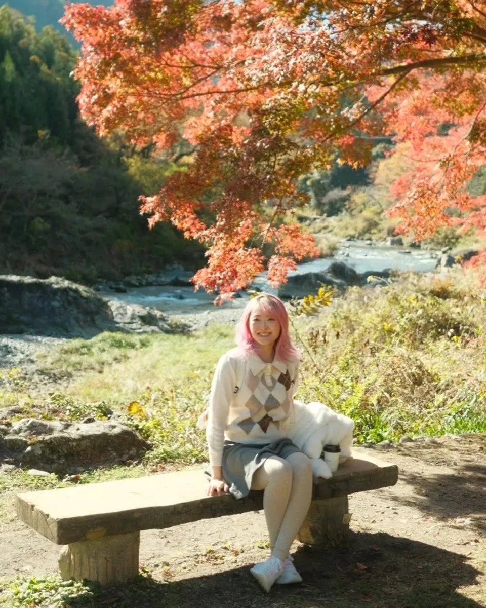 Xinni on a bench beside a river under red autumn maples