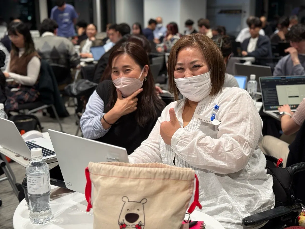 Two women in face masks at a front-row table with laptops open, one giving a thumbs-up, in a packed session where every attendee behind them is also on a laptop