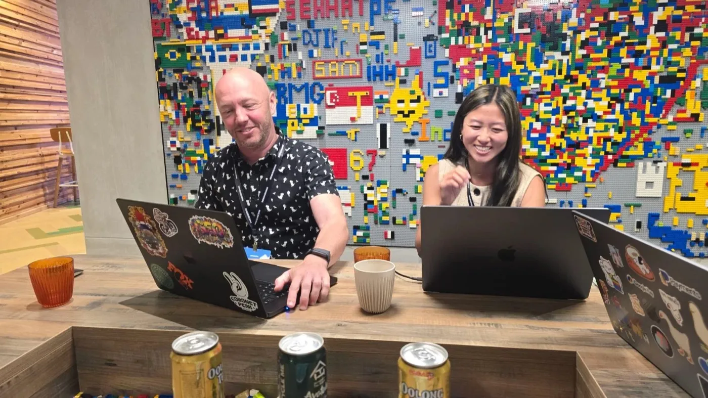 Two people laughing at their laptops in front of a large Lego wall