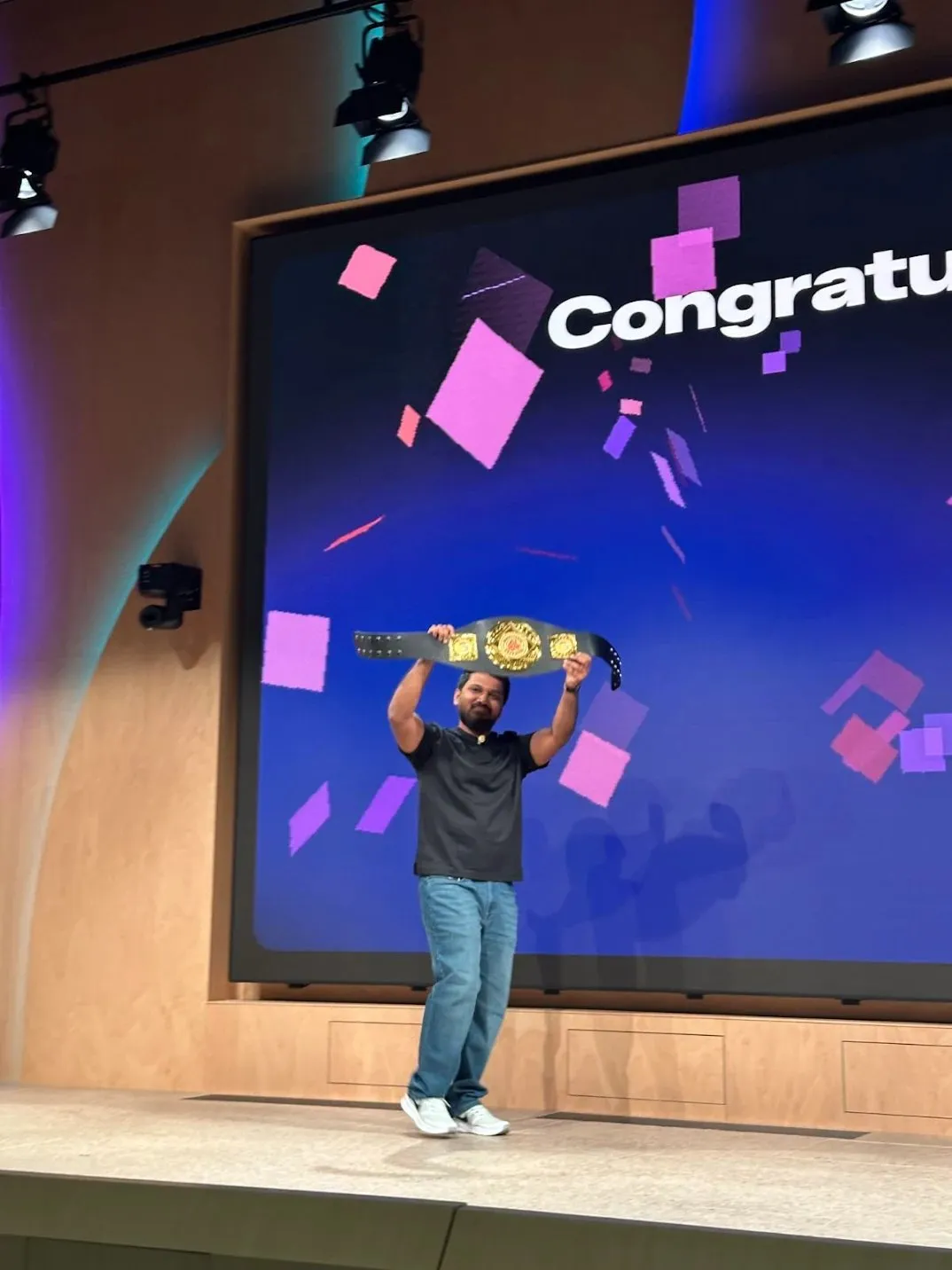 A contest winner on stage holding the championship belt overhead in front of a giant Congratulations screen with confetti
