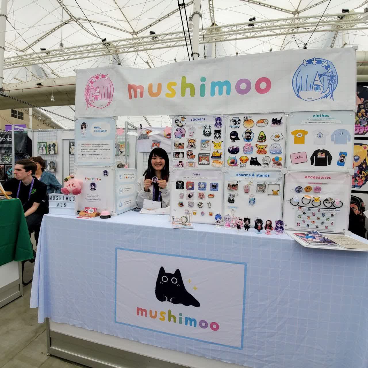 Xinni at her mushimoo market booth: a white banner with the mushimoo logo, walls of stickers, pins, charms, and clothes.