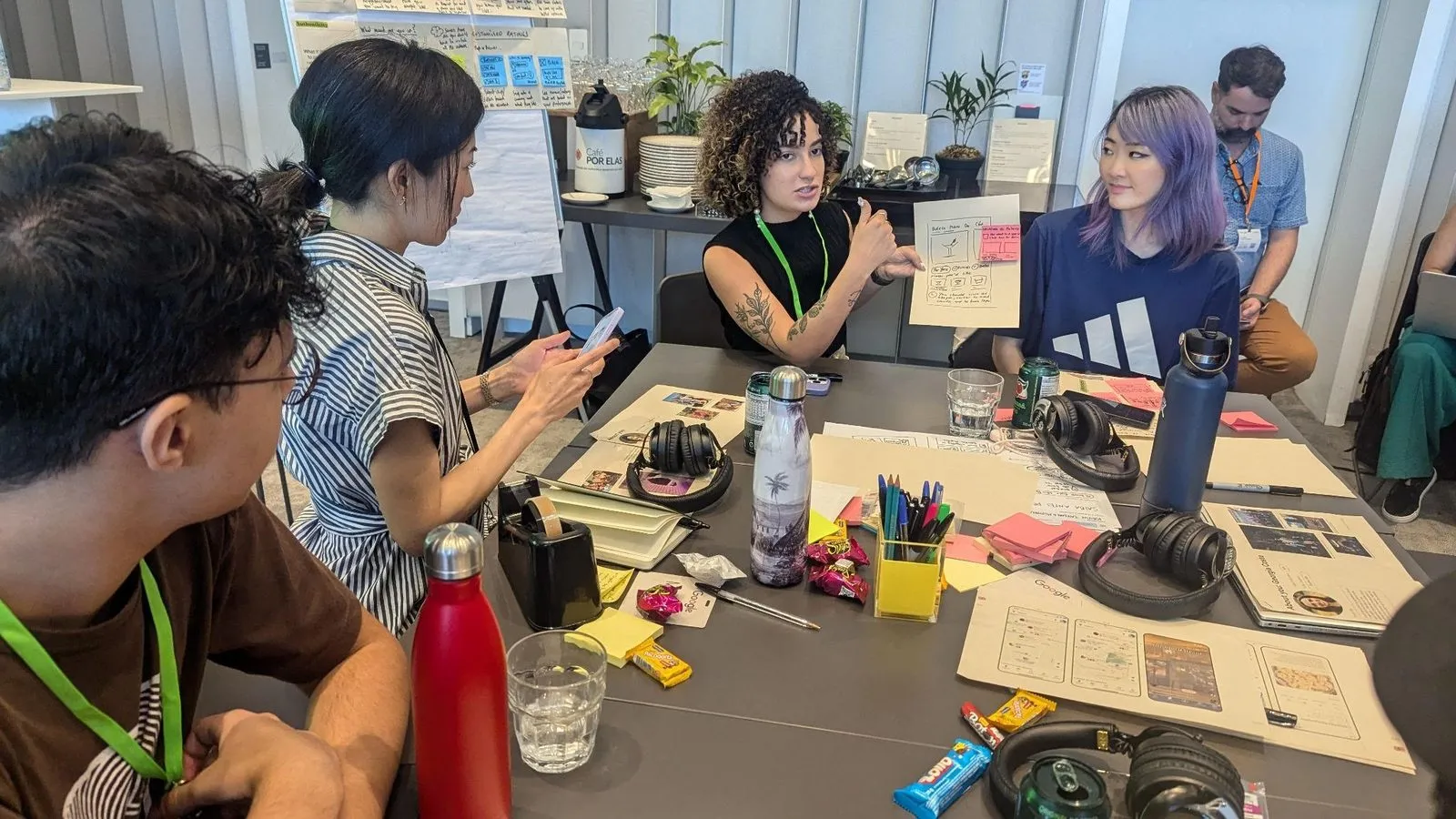Xinni at a table in the Sao Paulo co-creation workshop while a participant presents a sketched concept over printed screens and sticky notes