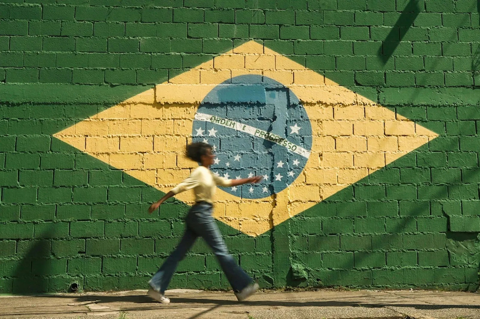 A person walking past a large Brazilian flag painted on a green brick wall, the yellow diamond and blue globe reading Ordem e Progresso