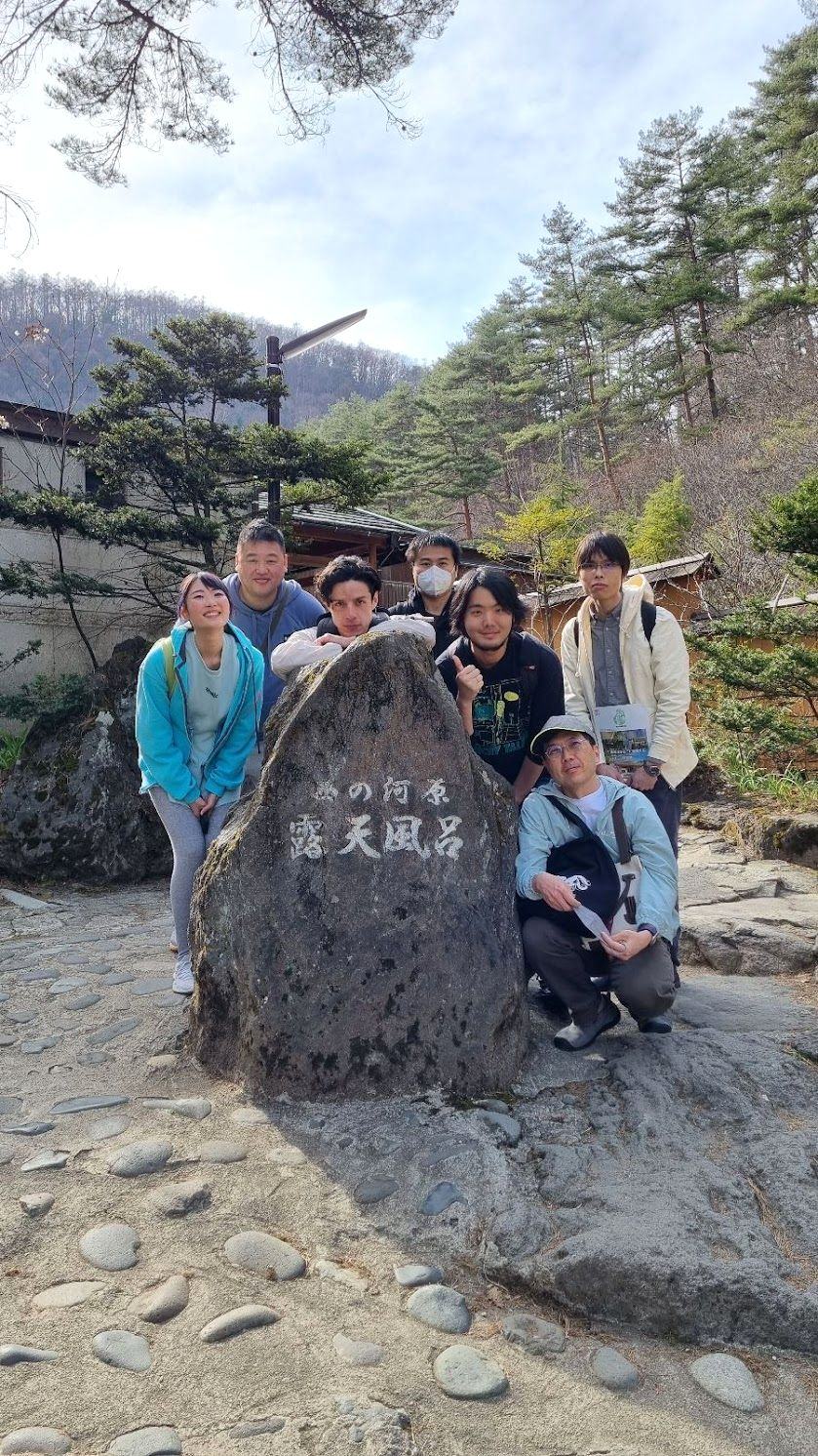 Seven members of the Google Travel Japan team crouched and standing around the stone marker for Kusatsu's Sainokawara open-air bath, pine hills behind them