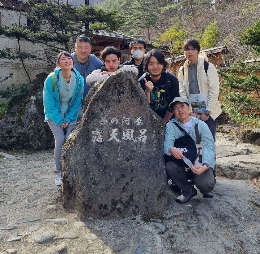 Seven members of the Google Travel Japan team crouched and standing around the stone marker for Kusatsu's Sainokawara open-air bath, pine hills behind them