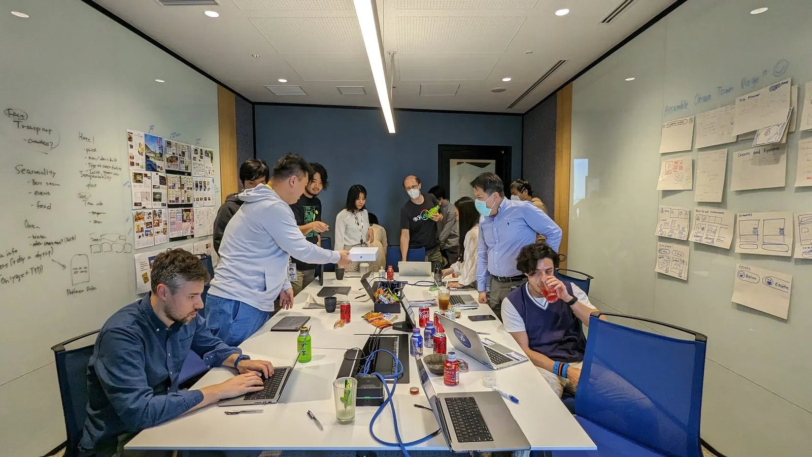 A dozen people mid-workshop around a long table of laptops and drinks; the left whiteboard breaks down transport, hotel, seasonality and onsen attributes, the right wall is covered in hand-drawn wireframes headed Assemble Onsen Town Package