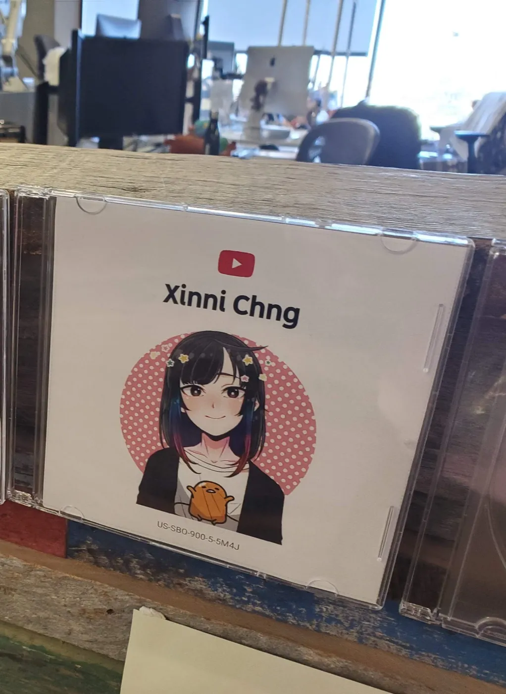 A CD jewel case on an office shelf printed with the YouTube logo, 'Xinni Chng', and an anime avatar of Xinni holding a small orange plush
