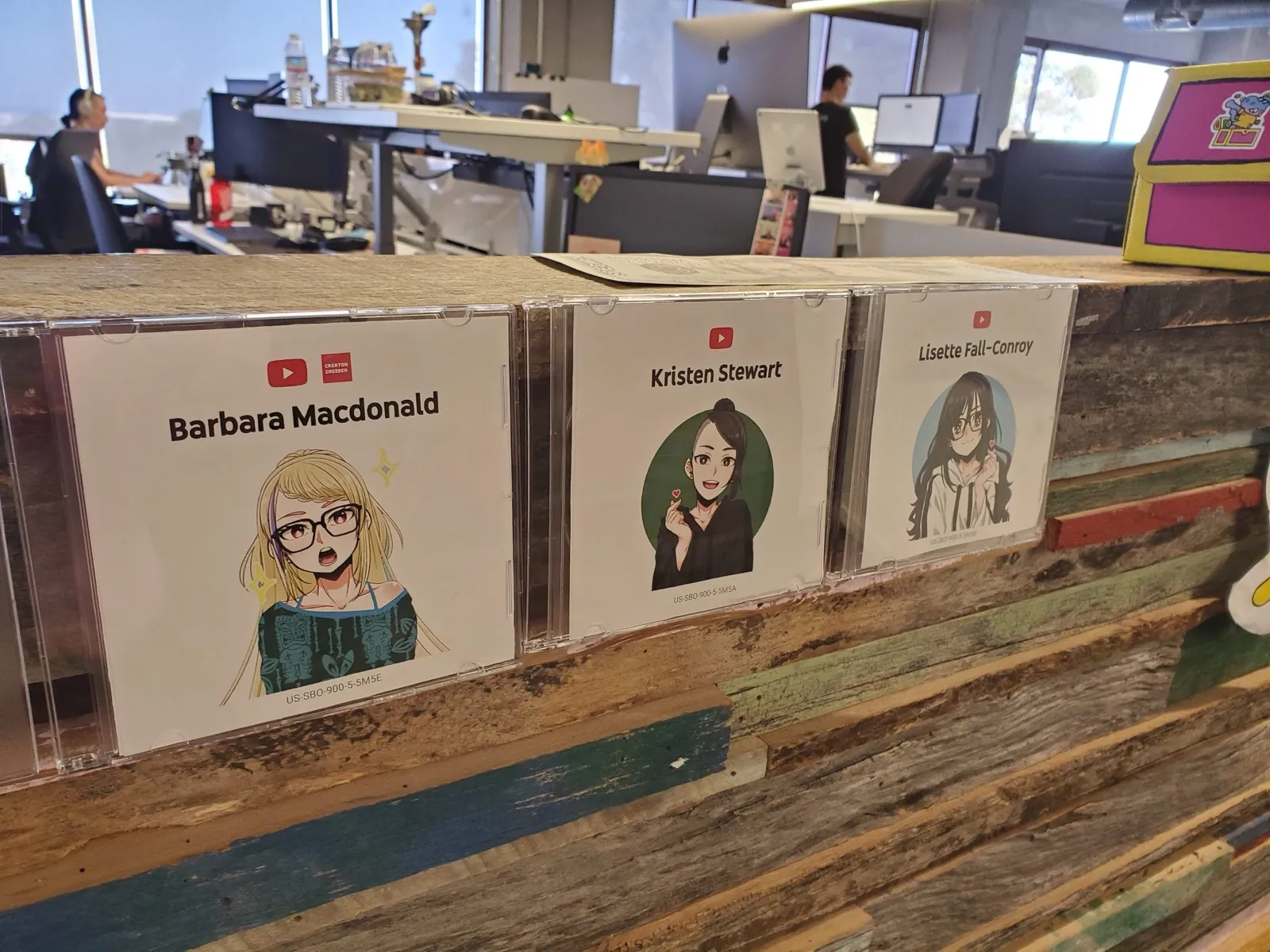 Three anime avatar badges in CD jewel cases on a reclaimed-wood shelf, labelled Barbara Macdonald, Kristen Stewart, and Lisette Fall-Conroy