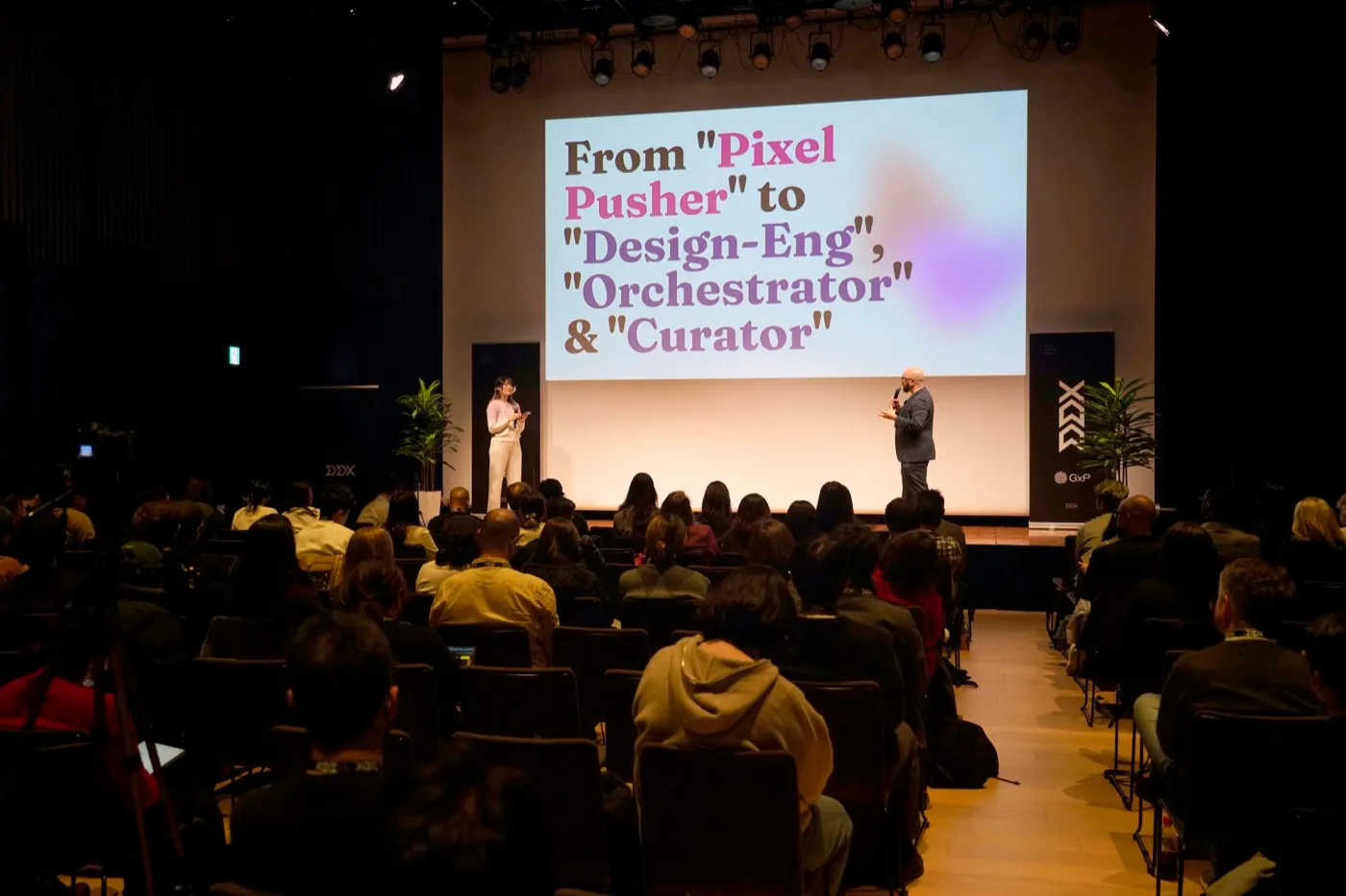 Xinni on stage at DDX Tokyo in front of a full auditorium, beside a slide reading From "Pixel Pusher" to "Design-Eng", "Orchestrator" & "Curator"