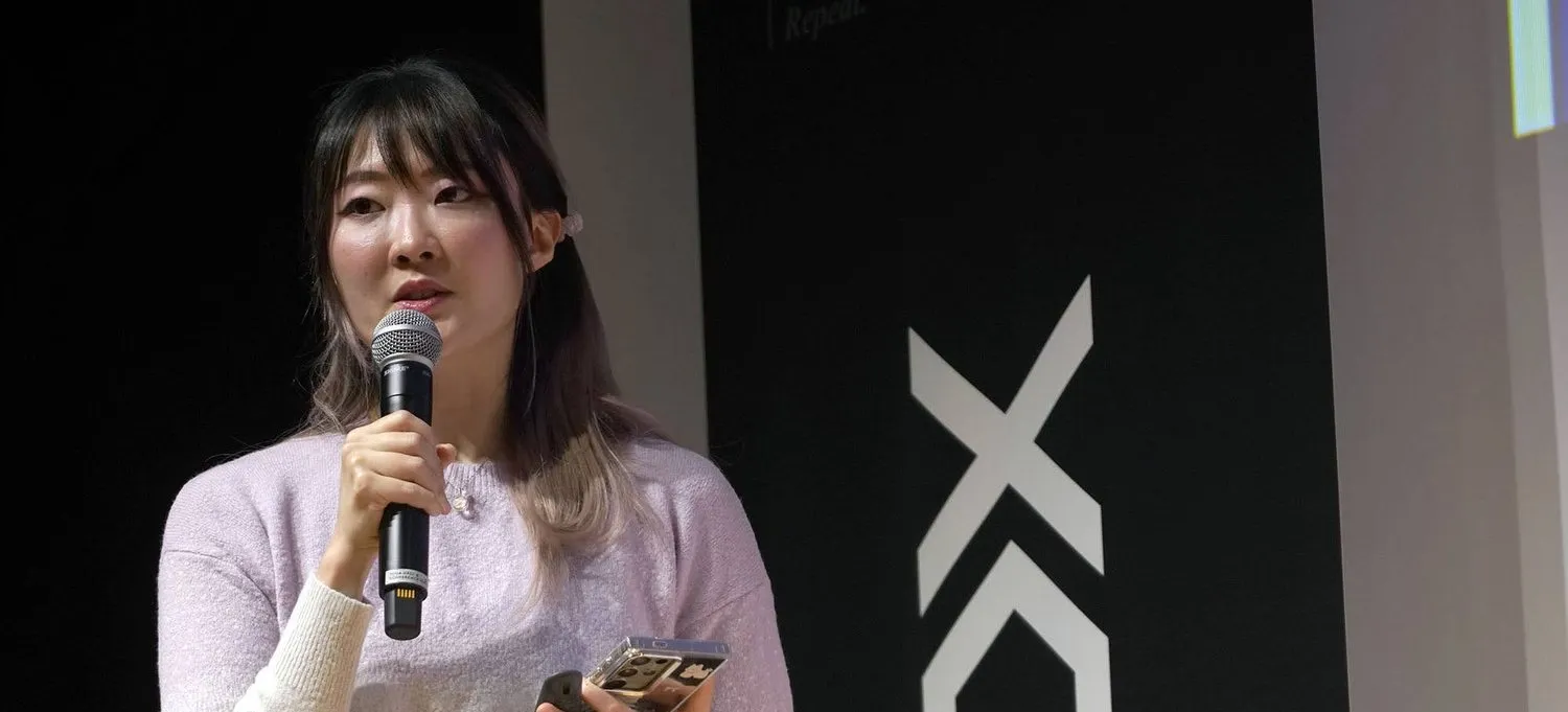 Xinni speaking at DDX Tokyo, microphone in one hand and her phone in the other, in front of a black banner carrying the DDX chevron mark