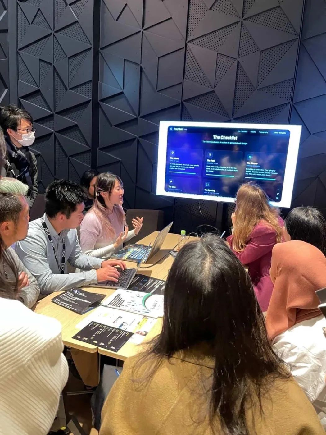 Xinni mid-sentence at a packed workshop table, gesturing at a wall screen showing a checklist of the commandments of modern AI-generated web design