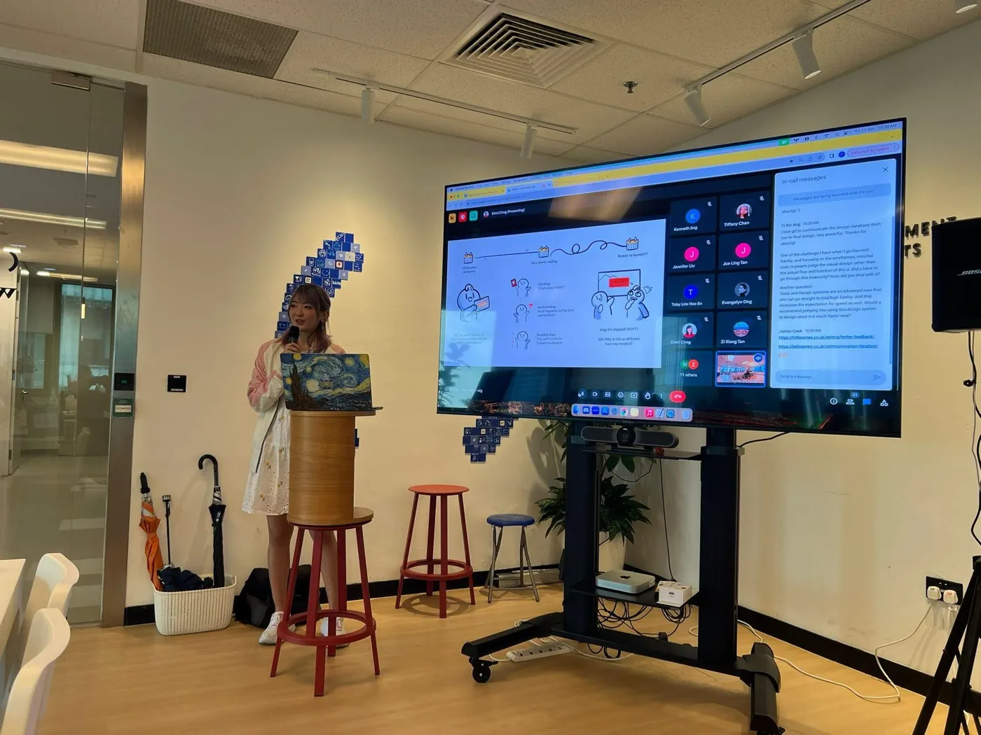 Xinni presenting at a podium to a hybrid room at OpenGov, the screen showing a comic strip of a design-to-engineering handoff next to a grid of remote attendees