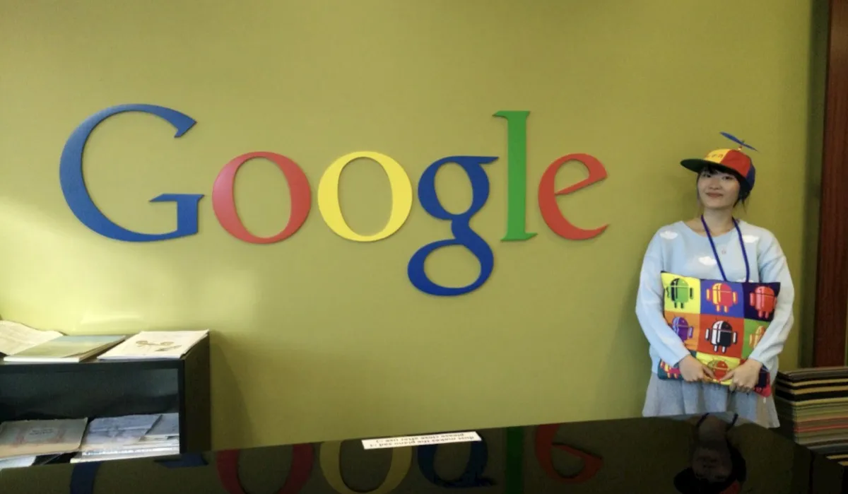Xinni in a propeller cap holding an Android cushion, standing beside the Google logo on an office wall